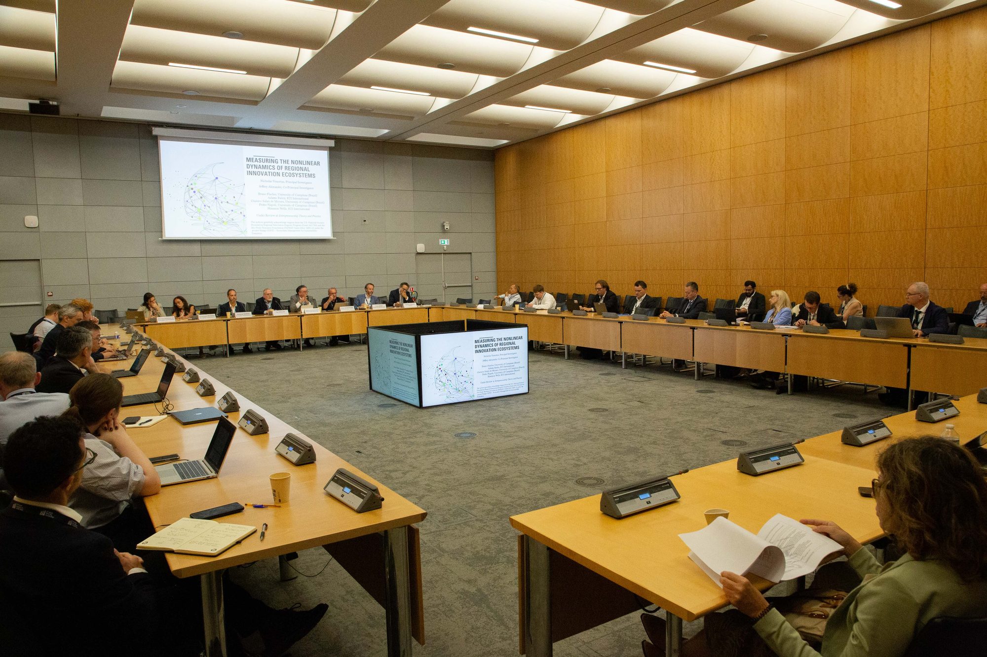 Participants seated around a square table arrangement during a parallel session, with a slide on regional innovation ecosystems on screen